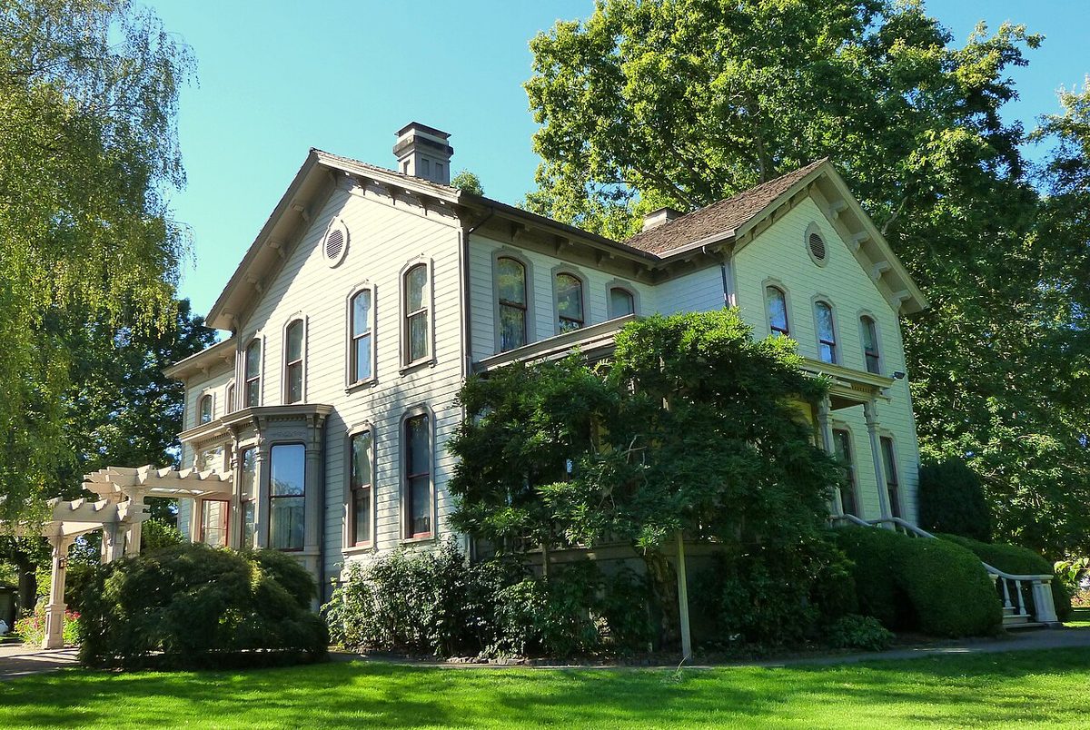 The cream-colored two-story Bush House in Salem, Oregon, framed by trees and a lawn