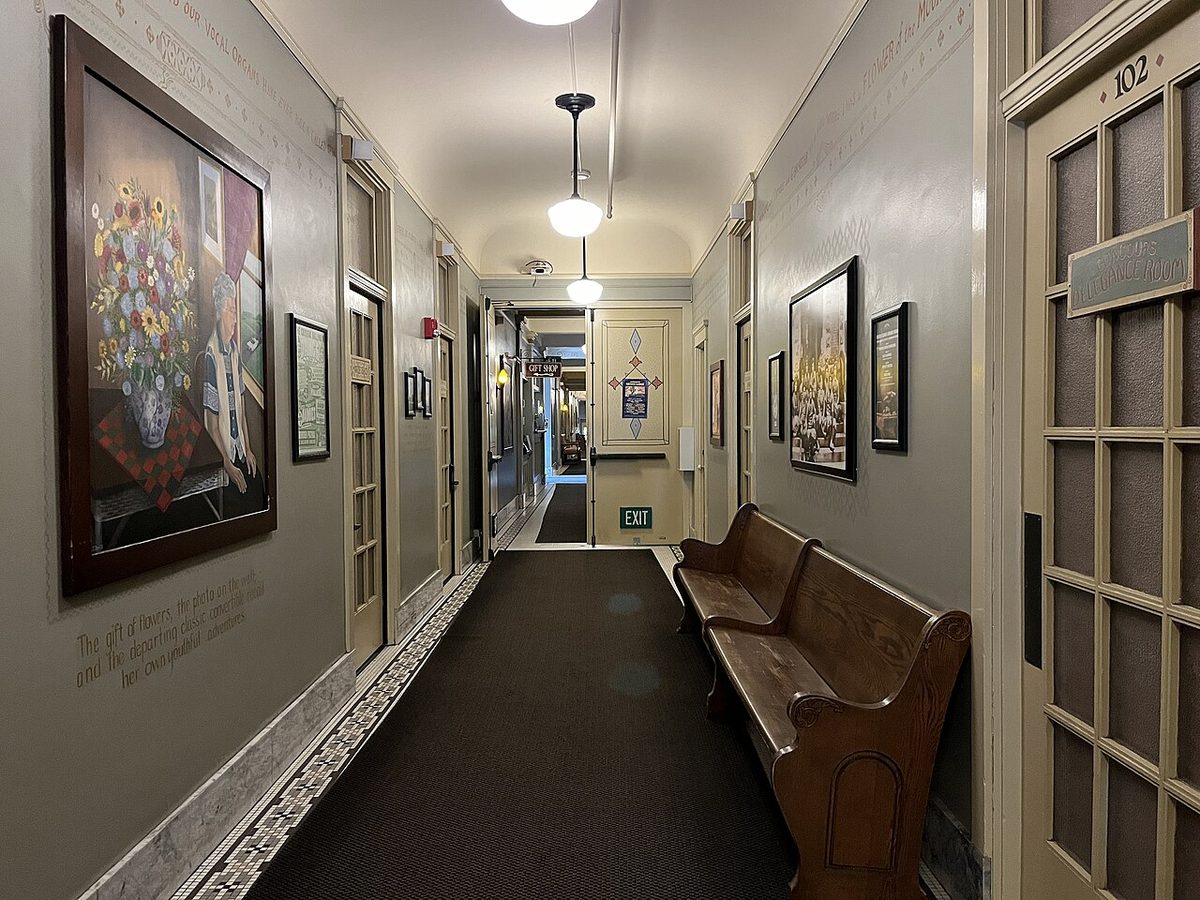 A long guest-room hallway inside McMenamins Grand Lodge with painted art panels on the walls and a wooden bench