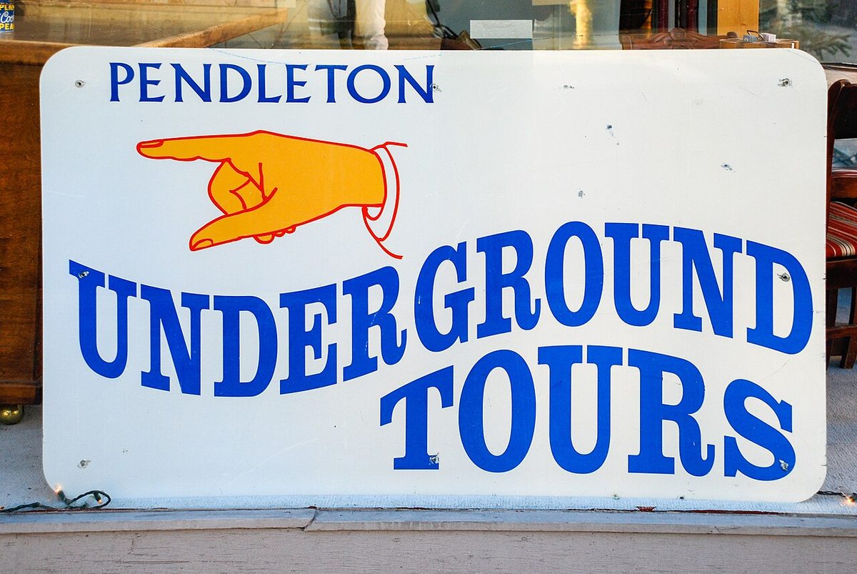 A white sign with a pointing hand reading Pendleton Underground Tours in blue letters