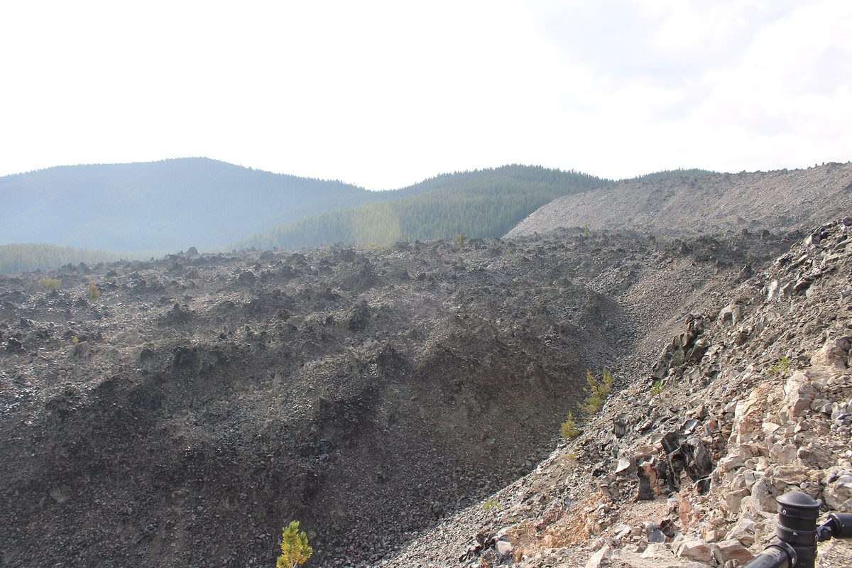 The steep, rubbly front of the Big Obsidian Flow rising above pine forest, with a trail post in the foreground