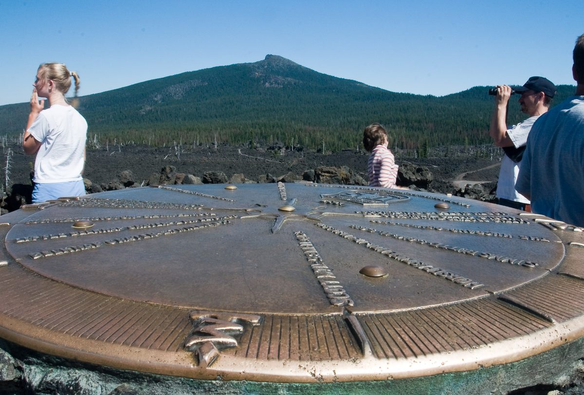 Visitors at the round bronze peak finder on the roof of Dee Wright Observatory, with a forested volcano behind