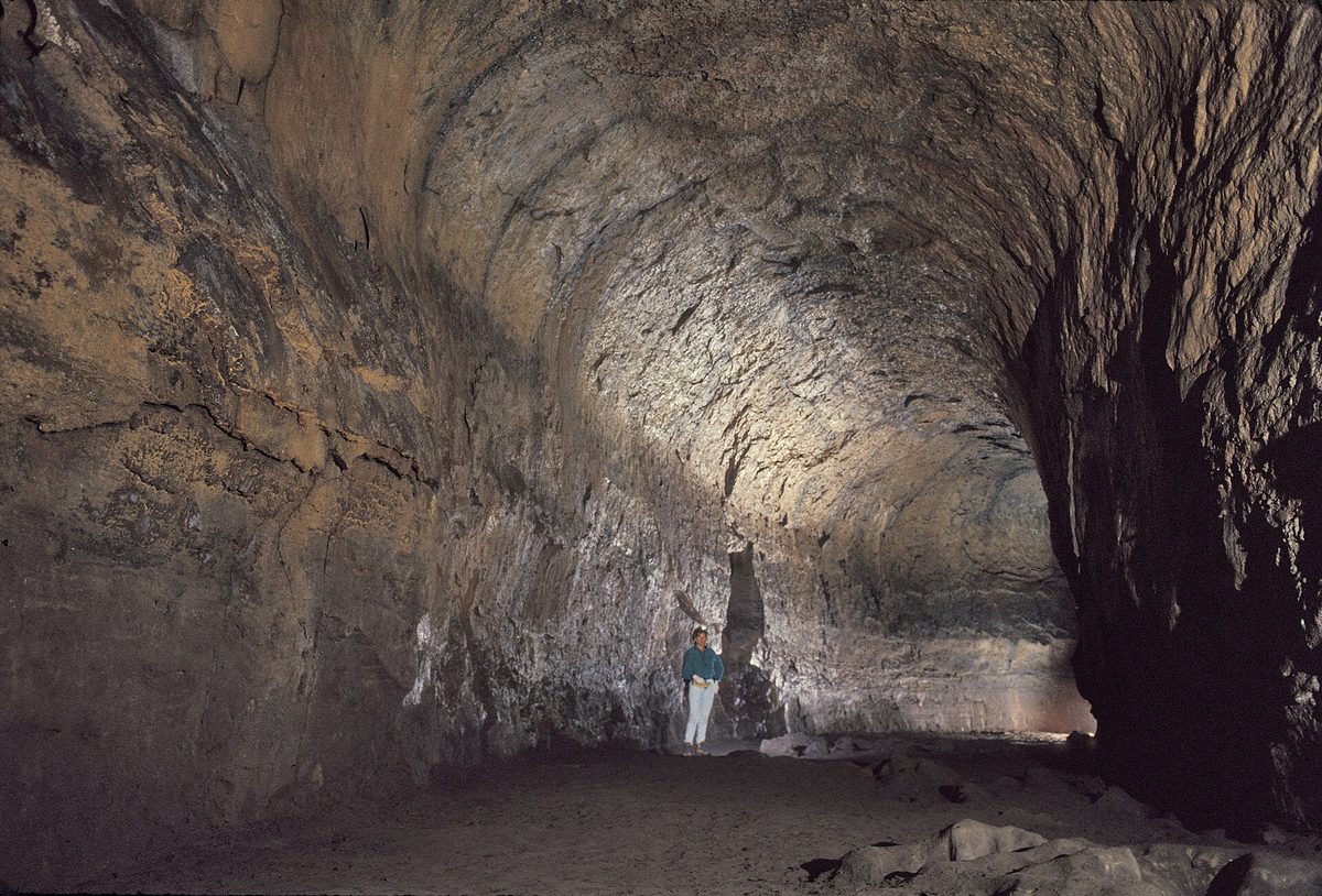 A wide, arched lava tube passage inside Lava River Cave lit by a lamp, with a person standing far down the tunnel