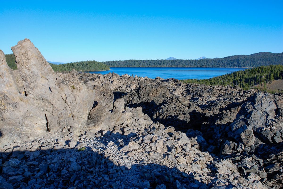Jagged black and gray obsidian and pumice of the Big Obsidian Flow, with blue Paulina Lake and forested ridges behind