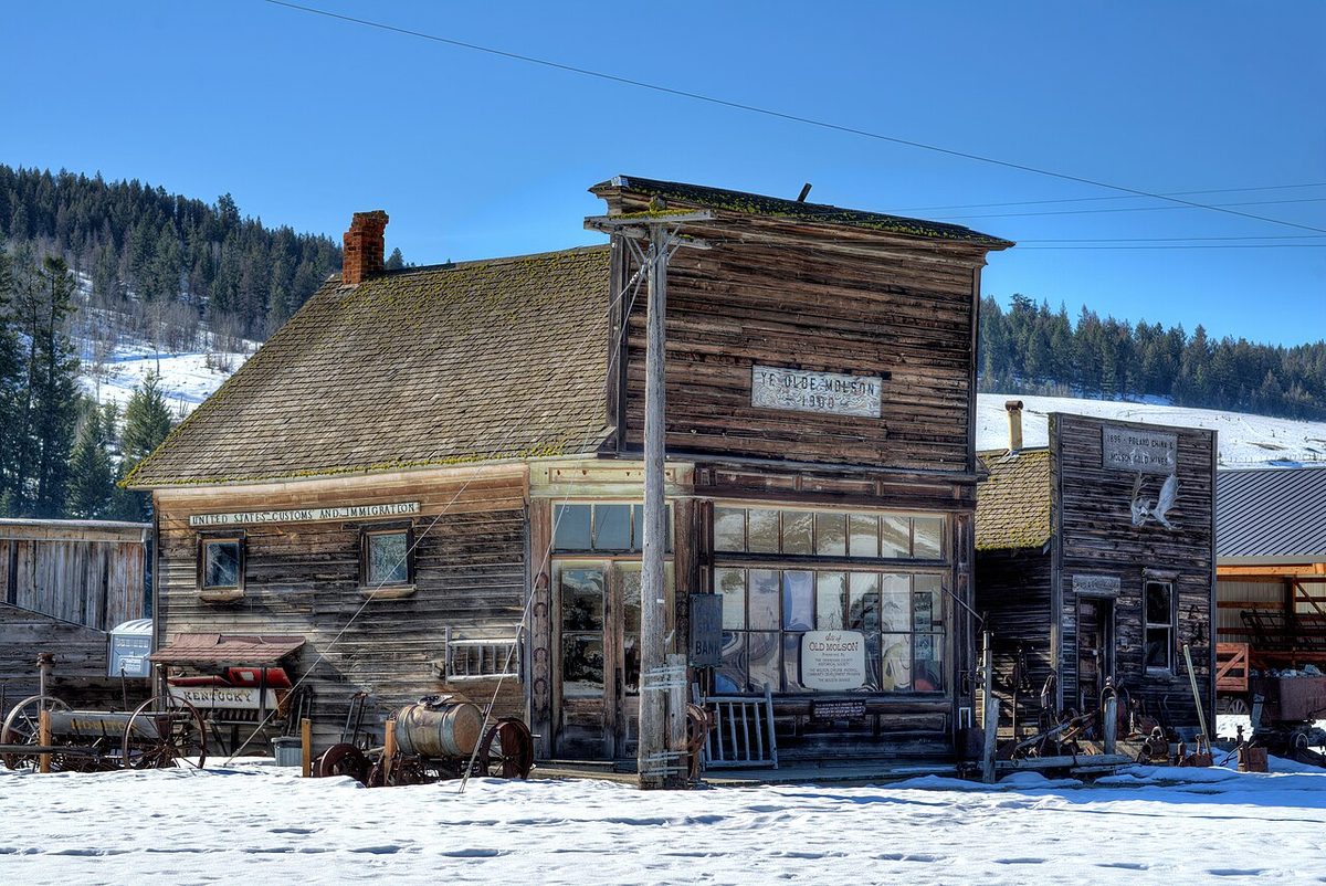 Weathered wooden false-front buildings in snow at Molson, with forested hills behind