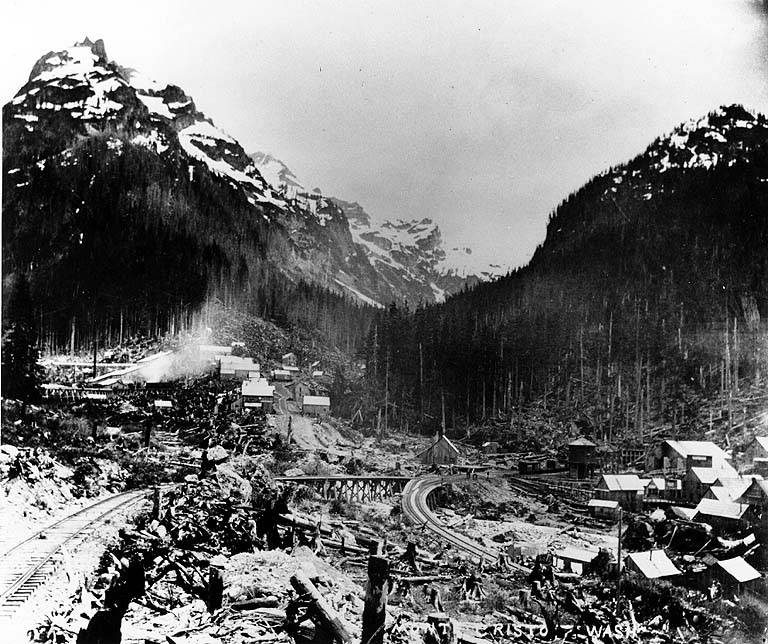 Black-and-white photo of the mining town of Monte Cristo in 1895, with buildings and rail yards below steep mountains