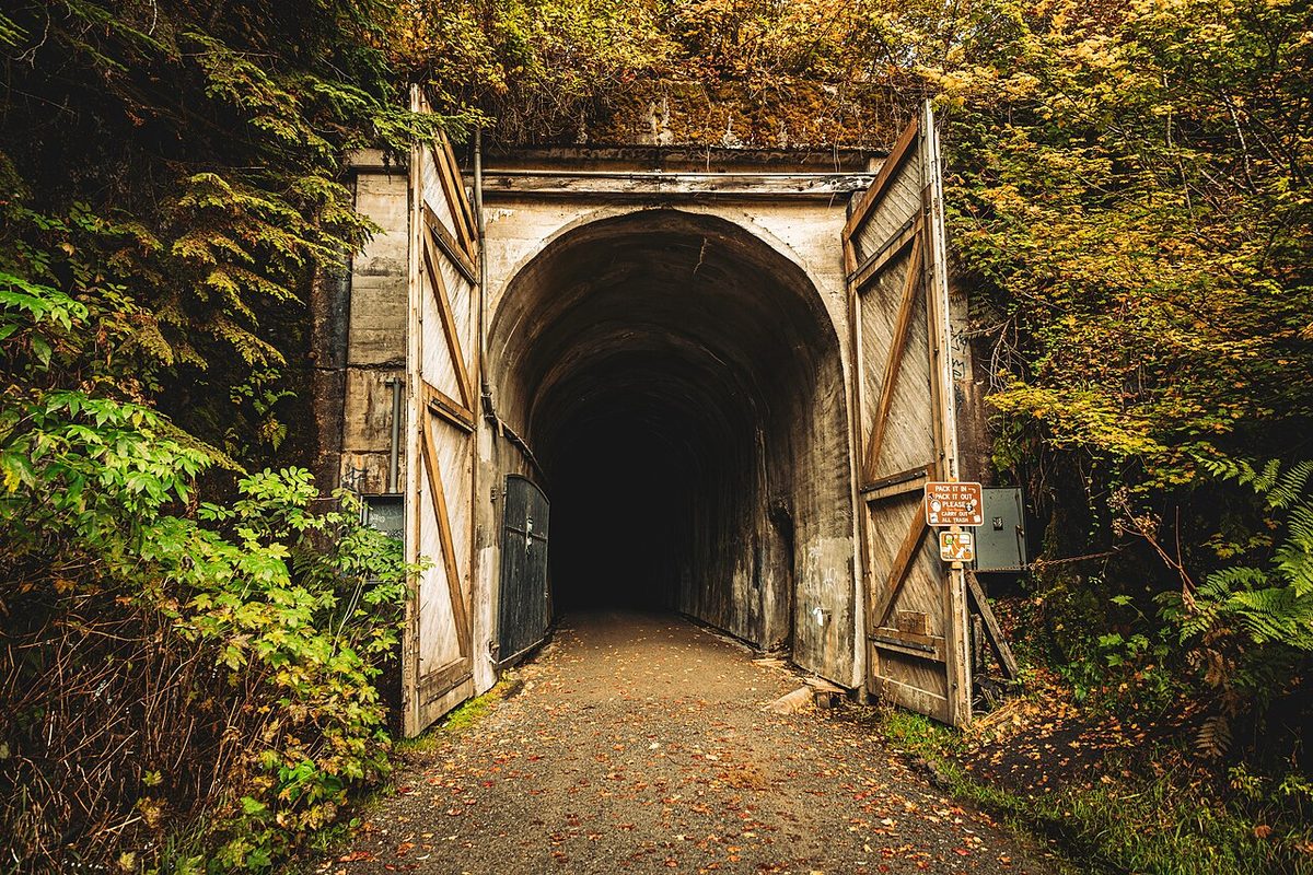 The arched concrete east portal of the Snoqualmie Tunnel, framed by fall leaves, with a gravel trail leading into the dark