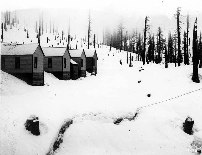 Black-and-white photo of snow-buried railroad buildings at Wellington with burned snags on the slope above