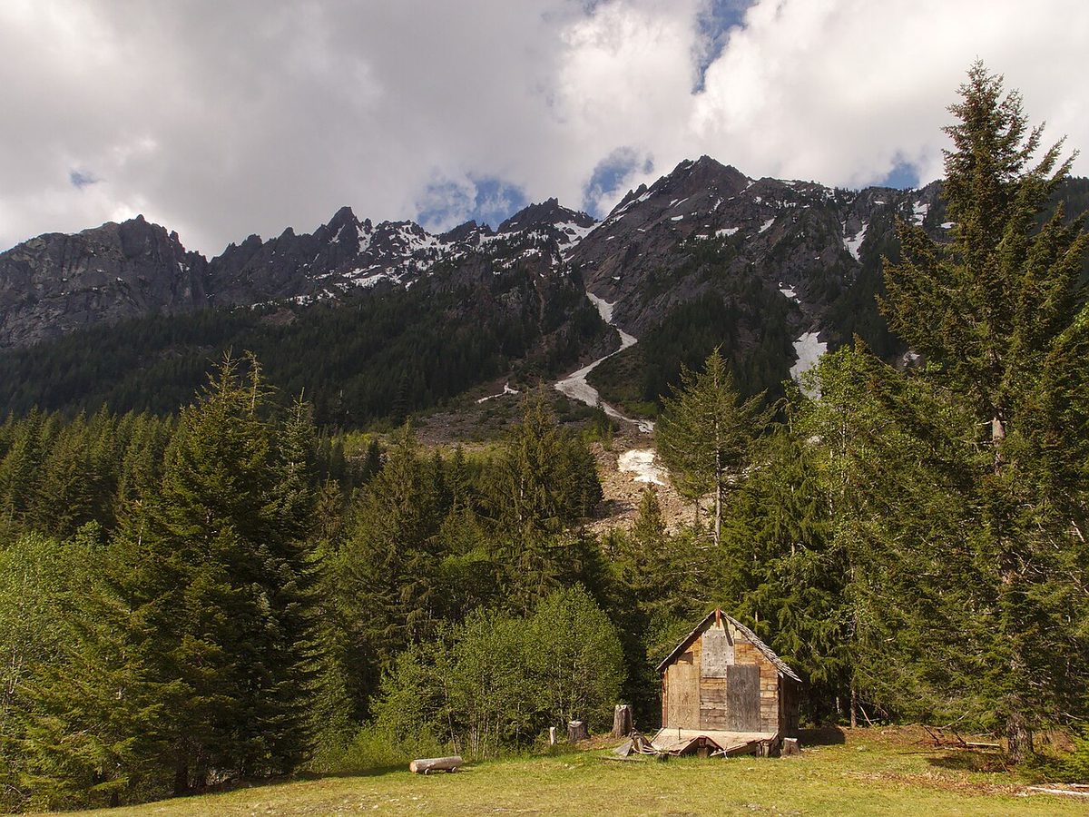 Weathered wooden cabins and foundations in a green clearing at the Monte Cristo townsite, with snowy peaks behind